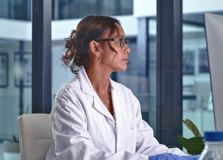 A woman in a lab coat with her hair up working on a desktop.