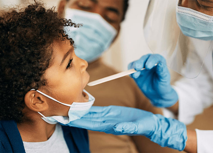 African American doctor with face mask examining boy's throat during a home visit.