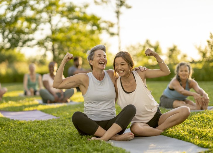 Two woman share a yoga mat as they lean in closely for a hug and flex their muscles. They are seated outdoors among their peers during a group fitness class. Each is dressed comfortably in athletic wear.