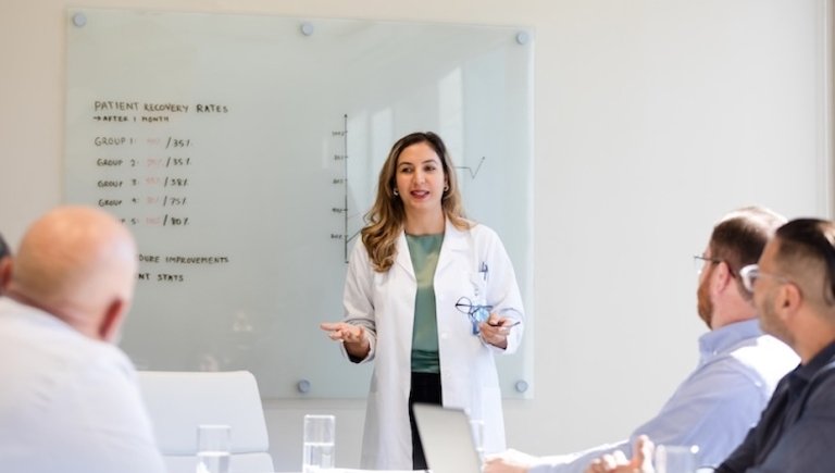 Doctor stands in front of white board speaking to colleagues. 