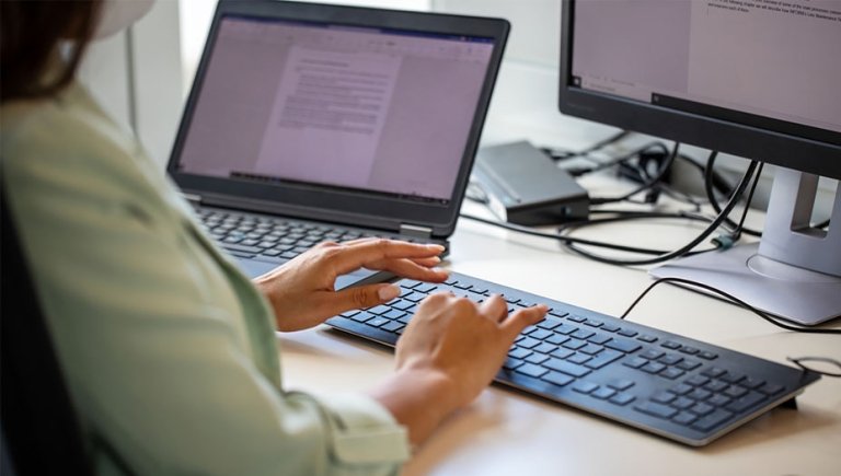 A person typing on a keyboard with a computer in the background. 
