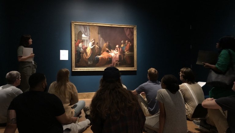 People sitting in a museum with a spotlight on a painting.