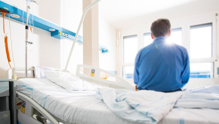 A hospital patient with back to camera sitting on a bed looking out the window.