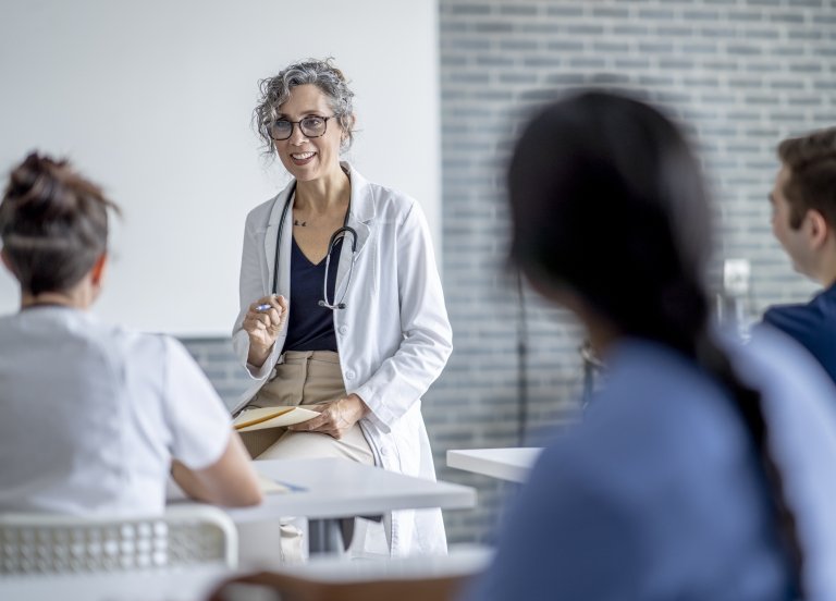 Medical educator wears a white coat and sits in front of students in a classroom.