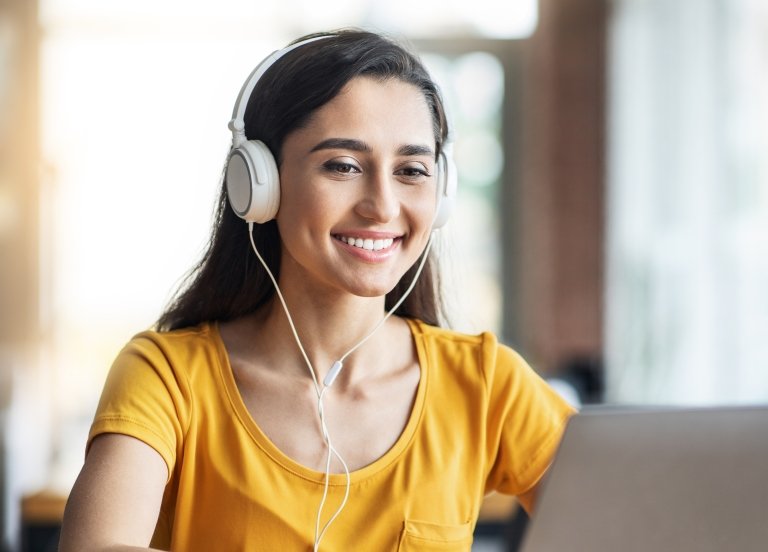 Young woman wearing a yellow shirt and headphones smiles while looking at her laptop.